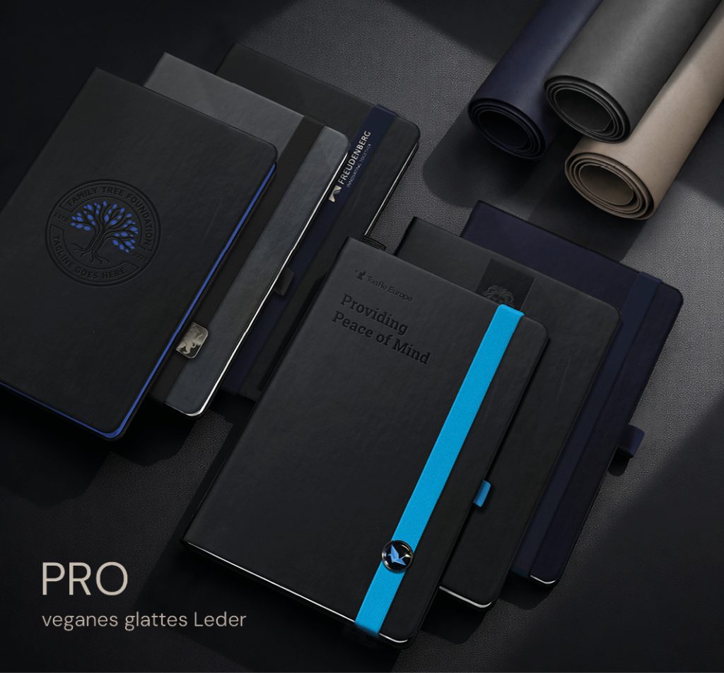 Hochwertige Lanybook® PRO Notizbücher aus veganem glattem Leder in dunklen Farbtönen, mit individueller Logo-Prägung, farbigem Verschlussband und Metall-Button, arrangiert auf einer edlen Materialoberfläche mit gerollten Covermaterialien im Hintergrund.