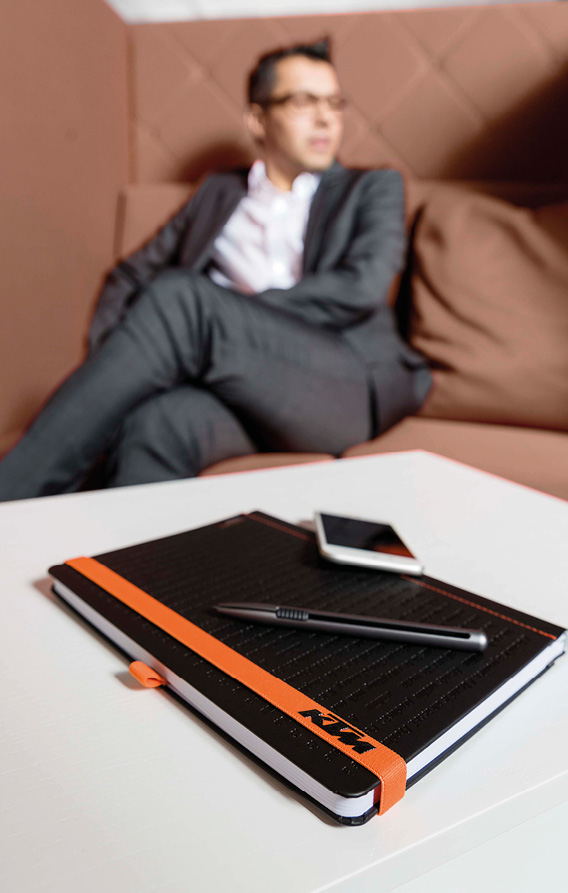 Notizbuch mit schwarzem Einband und orangefarbenem Gummiband liegt auf Tisch, im Vordergrund Stift und Smartphone, Business-Szene mit sitzender Person im Hintergrund