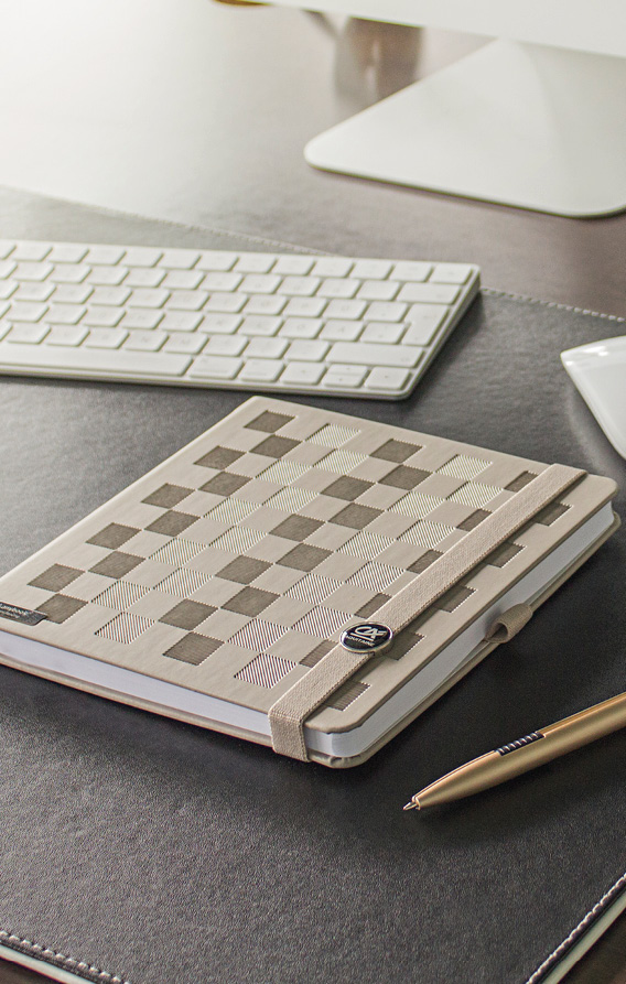 Beiges Notizbuch mit Karomuster liegt auf Schreibtisch neben Tastatur und Stift, elastisches Verschlussband mit Metallemblem sichtbar
