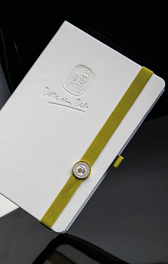 Weißes Notizbuch mit geprägtem Logo und grünem elastischem Verschlussband, hochwertig inszeniert auf dunklem Hintergrund