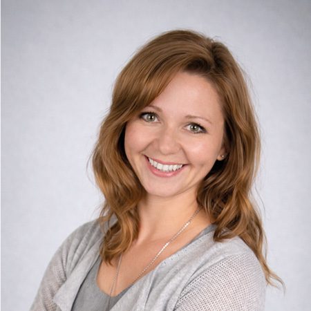 Porträt einer lächelnden Frau mit verschränkten Armen und grauer Kleidung vor neutralem Studiohintergrund, Mitarbeiterin eines Notizbuchherstellers