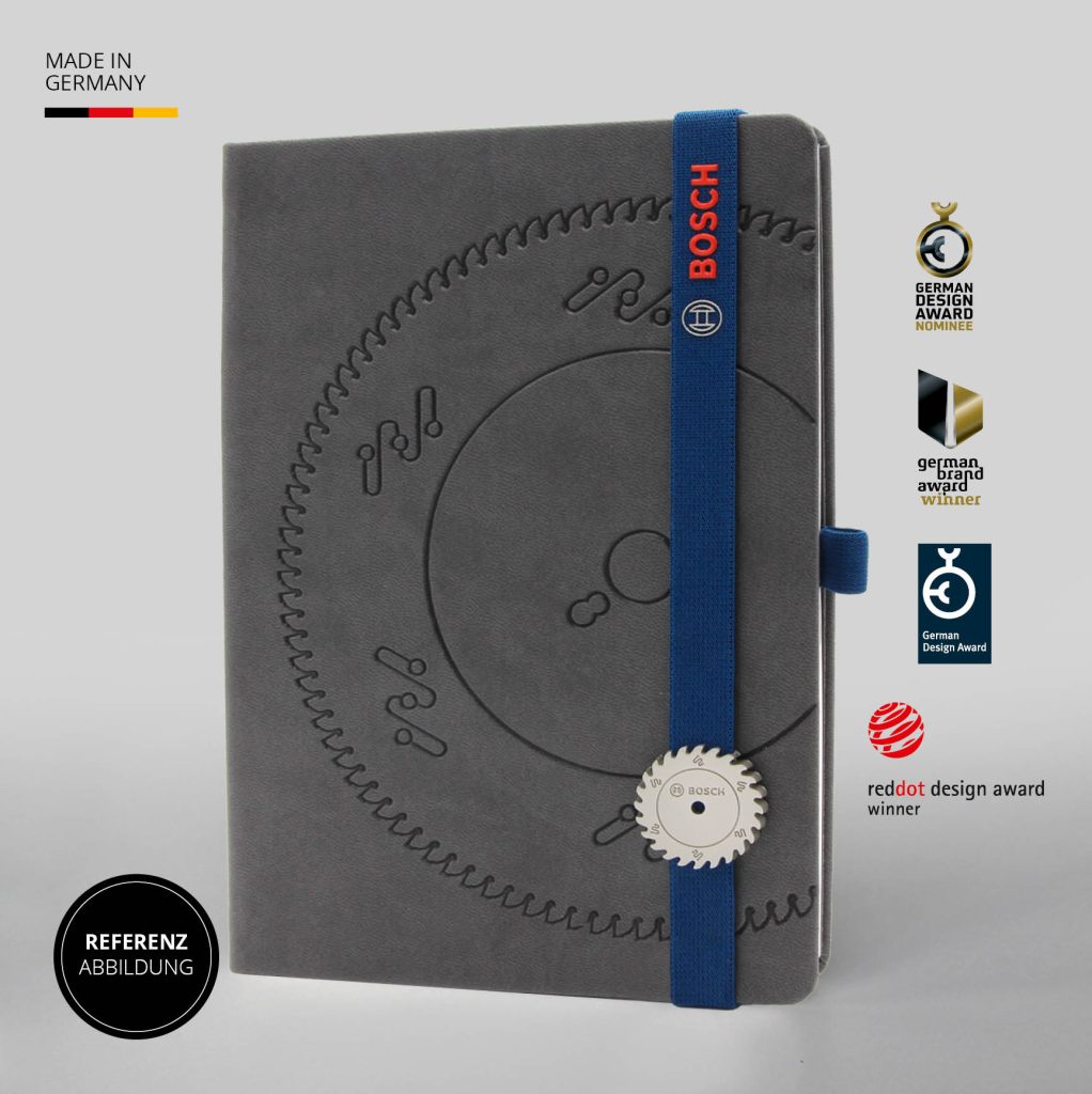 Graues Notizbuch mit großflächiger technischer Blindprägung, blauem Lanyband mit BOSCH Schriftzug und dekorativem Button, Referenzabbildung mit Design Awards