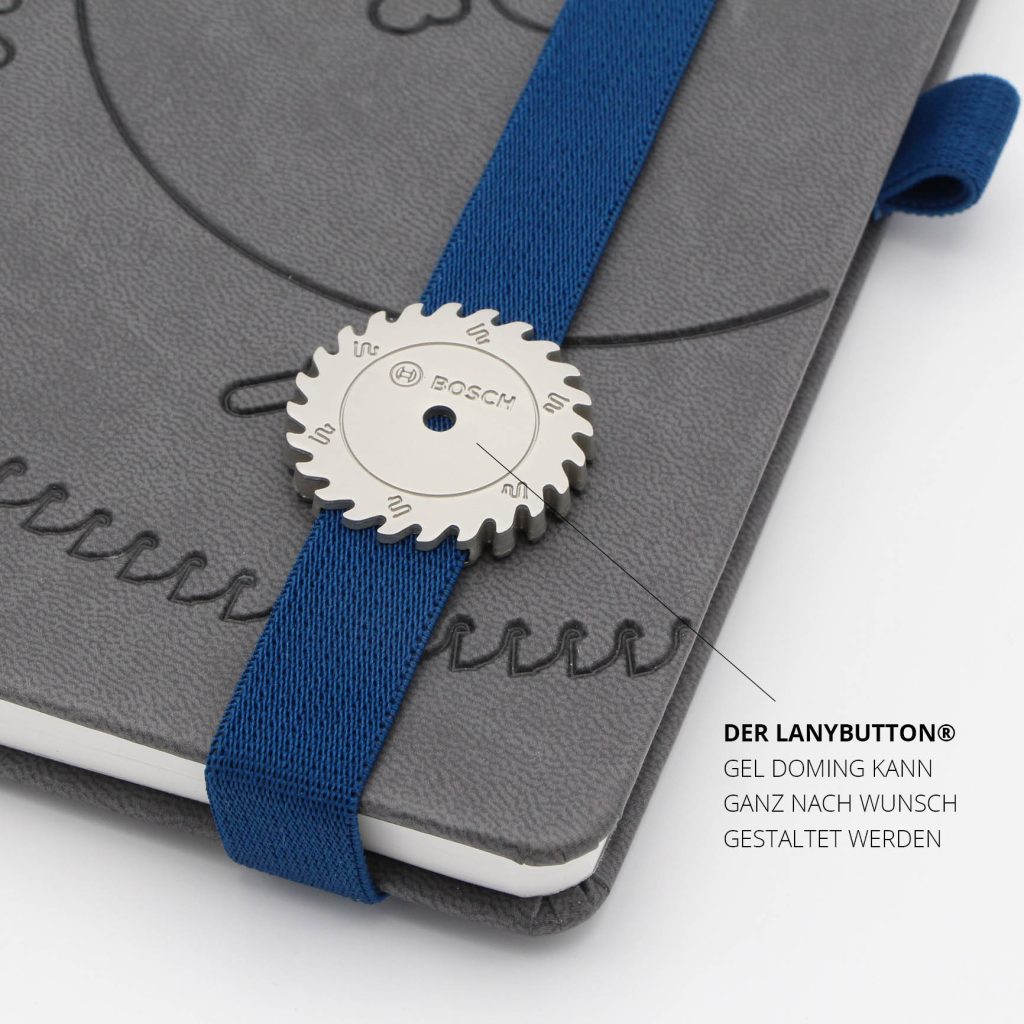 Detailansicht des runden LanyButtons mit Bosch Zahnraddesign auf blauem Lanyband und grauem Notizbuchcover