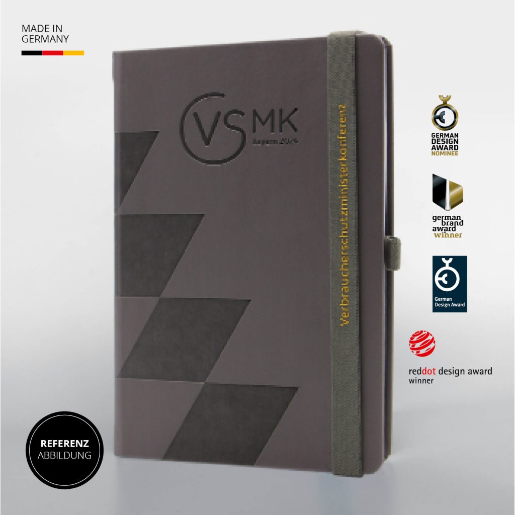 Dunkelgraues Notizbuch mit geometrischem Muster und VSMK Logo, grauem Lanyband mit gelbem Schriftzug, Referenzabbildung Made in Germany mit Auszeichnungen
