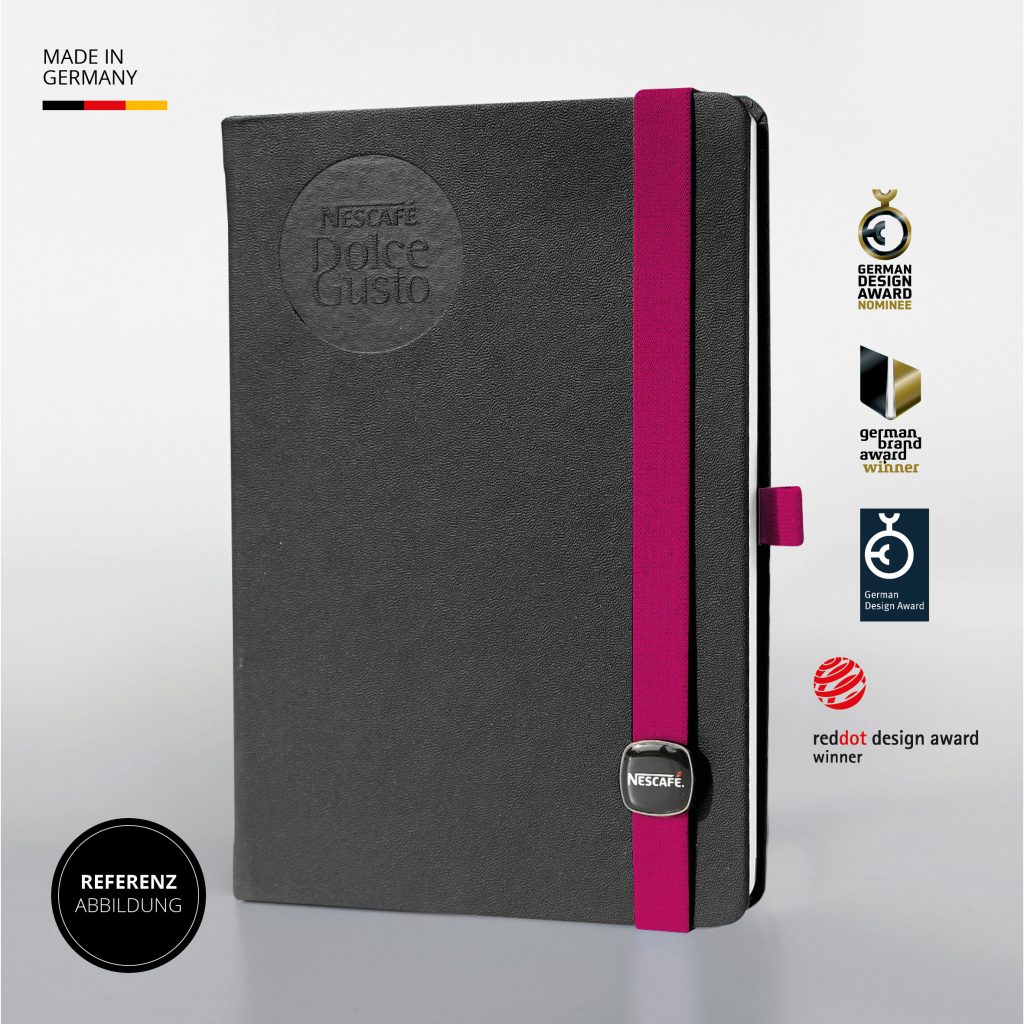 Schwarzes Notizbuch mit runder Nescafé Dolce Gusto Blindprägung, himbeerfarbenem Lanyband mit Logo Button, Referenzabbildung Made in Germany mit Design Awards