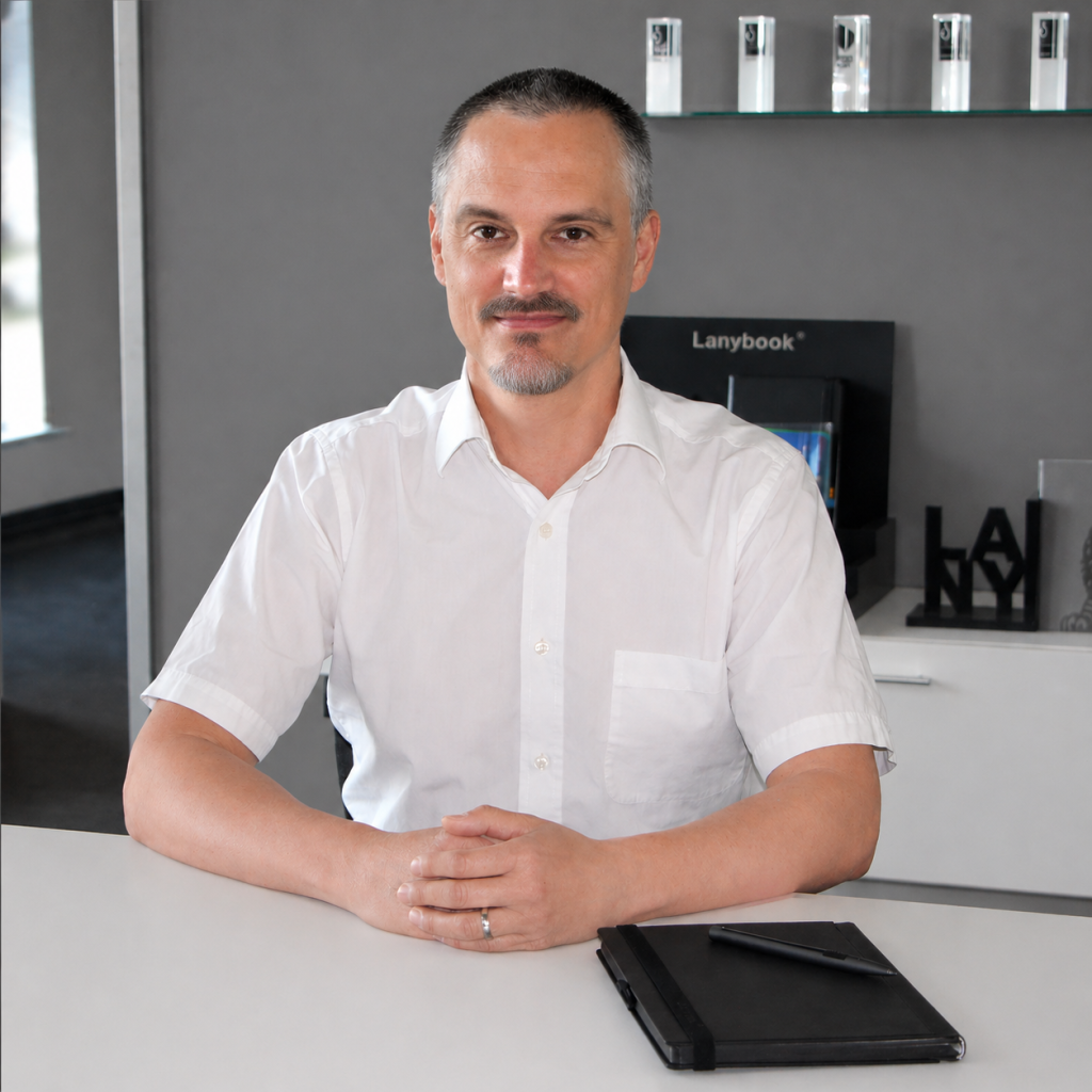 Vertriebsleiter im weißen Hemd sitzt an einem Besprechungstisch im modernen Büro, die Hände gefaltet, vor sich ein schwarzes Notizbuch mit Stift. Im Hintergrund sind eine Lanybook® Präsentationsfläche, Regale mit Auszeichnungen und dezente Büroelemente zu sehen.