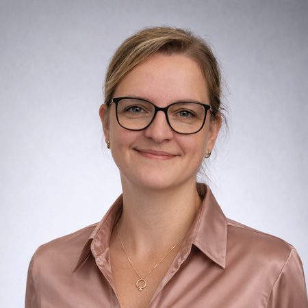 Porträt einer Frau mit Brille und rosafarbener Bluse vor neutralem grauem Hintergrund, Mitarbeiterin im Produktmanagement eines Notizbuchherstellers